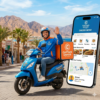 Captain Dahab Delivery App rider on a blue scooter in Dahab with the official orange logo, mountains, and Red Sea background